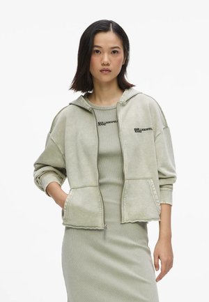 Frau trägt einen hellgrünen Kapuzenpullover mit Reißverschluss und ein passendes ärmelloses Kleid, beide mit dem "Karl Lagerfeld Jeans"-Logo auf der Brust.