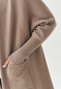 Cardigan en tricot beige avec des poignets côtelés et une poche. Le tissu semble doux, avec une texture lisse et un design décontracté.
