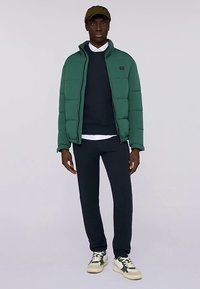 Zielona kurtka puchowa na zamek, noszona na granatowym swetrze, w połączeniu z ciemnymi spodniami dresowymi i białymi trampkami. Czapka dopełnia stylizację.