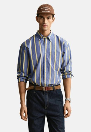 Mann trägt braune Mütze, blau-gelb gestreiftes Hemd mit Knöpfen und hochgekrempelten Ärmeln, braunen Gürtel, dunkle Jeans und silberne Armbanduhr.