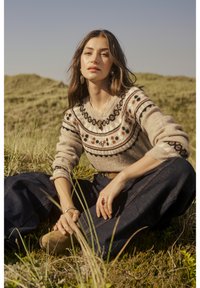 Bežs adīts džemperis ar aplīšiem melnā un brūnā krāsā, apvienots ar tumšiem plašiem džinsiem. Dabas vide ar zāli priekšplānā.