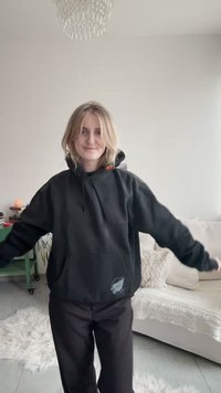 Fekete pulóver elülső zsebbel, húzózsinóros kapucnival és jobb alsó sarokban lévő logóval. Fekete nadrággal viselve, világos beltéri környezetben.