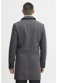 Manteau en laine grise avec une coupe ajustée, col cranté et poignets boutonnés. Le dos présente un design épuré et droit, avec un tissu texturé.
