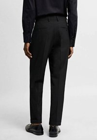 Czarne, dopasowane spodnie garniturowe o smukłym kroju, z gładką fakturą, dwoma tylnymi kieszeniami i delikatnie podwiniętymi nogawkami u dołu, w zestawieniu z czarnymi butami.