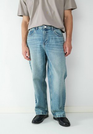 Ausgebleichte blaue Jeans mit lockerer Passform, gesteppten Details, fünf Taschen und einem Knopfverschluss. Kombiniert mit schwarzen Schuhen und einem grauen T-Shirt.