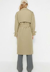 Osoba w beżowym trenczu, szarych spodniach i beżowej czapce baseballowej, stoi przodem do białej ściany, całkowicie widoczna od tyłu.