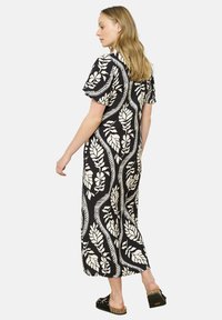 Abito maxi nero con motivo di foglie bianche, maniche corte, vestibilità ampia e texture liscia. Indossato con sandali neri con dettagli dorati.
