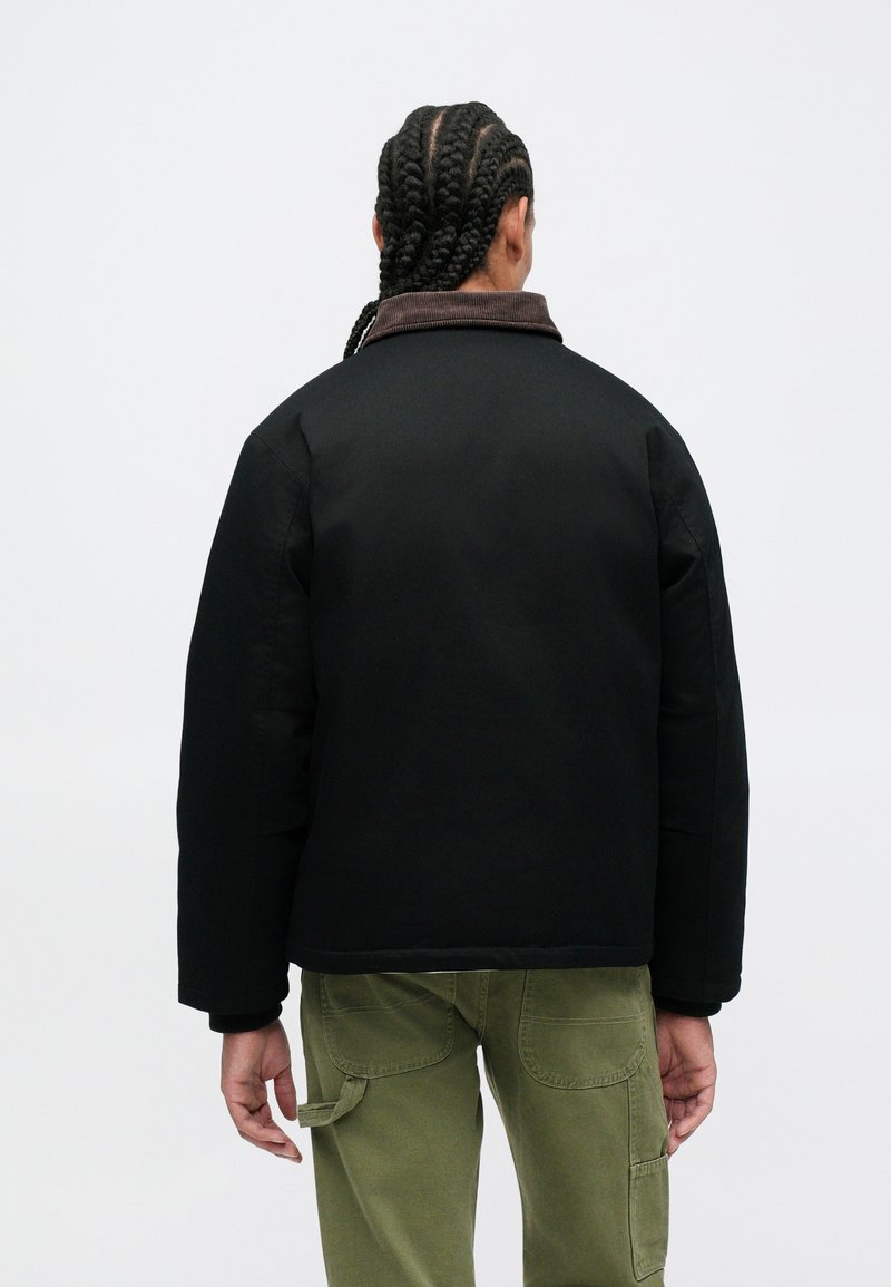 Persona con capelli intrecciati di spalle, che indossa una giacca nera con colletto marrone e pantaloni cargo verdi, in un contesto semplice.