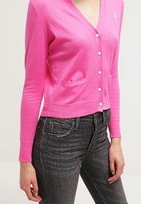 Kvinna klädd i en klarrosa knappad kofta med små fickor och mörkgrå tajta jeans, stående mot en enfärgad bakgrund.