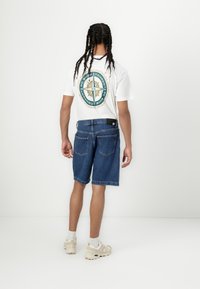 T-shirt blanc avec un graphique de boussole au dos, associé à un short en denim. Le short est bleu, arrive au genou, avec un ourlet brut et des poches arrière.