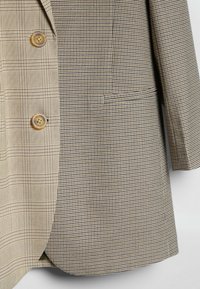 Blazer à carreaux bicolore avec un grand motif à carreaux beige sur le côté gauche et un petit motif pied-de-poule sur le côté droit, avec deux boutons marron.