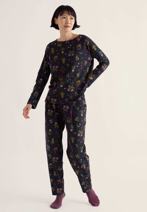 Femme debout portant un haut à manches longues et un pantalon assortis à motif floral foncé avec des chaussettes violettes sur un fond clair uni.