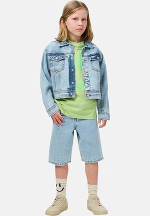 Giacca di jeans azzurro chiaro, t-shirt grafica verde e pantaloncini blu. Sneakers beige e calzini decorati con dettaglio di una faccina sorridente.