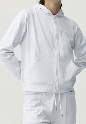 Person wearing a white zip-up hoodie with front pockets and matching white drawstring pants, standing with hands on hips.