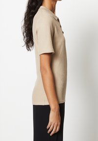 Beige ribbet polo t-shirt med kortærmer, der har tre knapper ved halsen og en tætsiddende pasform. Teksturen er glat og blød.