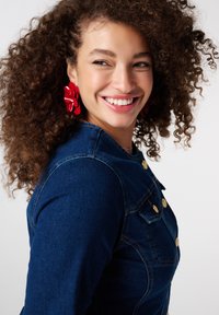 Vrouw met krullend haar, die rode bloemoorbellen en een donkerblauw spijkerjasje draagt, glimlacht en kijkt opzij tegen een witte achtergrond.