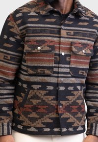 Wollen jas met een geometrisch patroon in tinten zwart, bruin en rood. Het heeft een knoopsluiting aan de voorkant, twee borstzakken en een kraag.