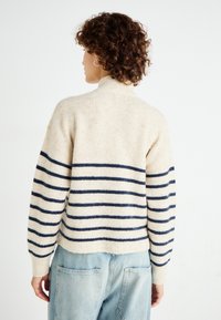 Knit sweater with a high collar, cream color featuring navy horizontal stripes, loose fit, and ribbed cuffs, paired with light blue jeans.