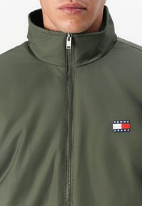 Olivengrønn zip-jakke med høy krage og sølvglider. Har et lite logo-patch på venstre bryst, laget av glatt stoff.