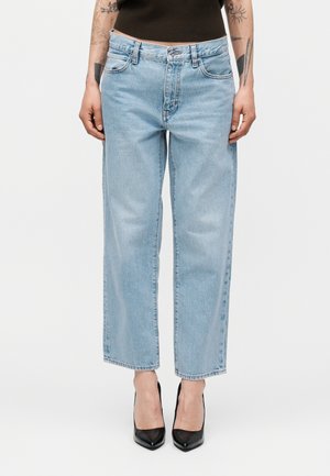 Személy világoskék egyenes szárú farmernadrágot, fekete hegyes orrú magassarkú cipőt és fekete ujjatlan felsőt visel, fehér háttér előtt állva.