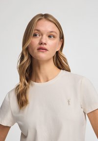 Katoenen T-shirt in lichtbeige met een ronde halslijn, korte mouwen en een klein goudkleurig logo op de linkerborst. Gladde textuur.