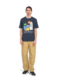 Marineblauw katoenen T-shirt met een grafische print van palmbomen, een gebouw en een zon; gecombineerd met kaki cargo pants en zwarte sneakers.