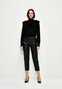 Zwarte coltrui met oversized schouders en een subtiel hartpatroon, gecombineerd met glanzende zwarte leren cropped broeken en zwarte hakken.