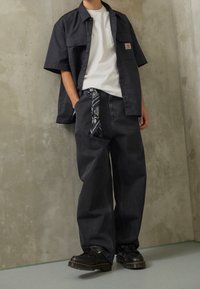 Dunkelgraue, locker sitzende Jeans mit einem am Bund gebundenen Bandana, kombiniert mit einem weißen T-Shirt und einem schwarzen kurzärmeligen Hemd mit Knöpfen.