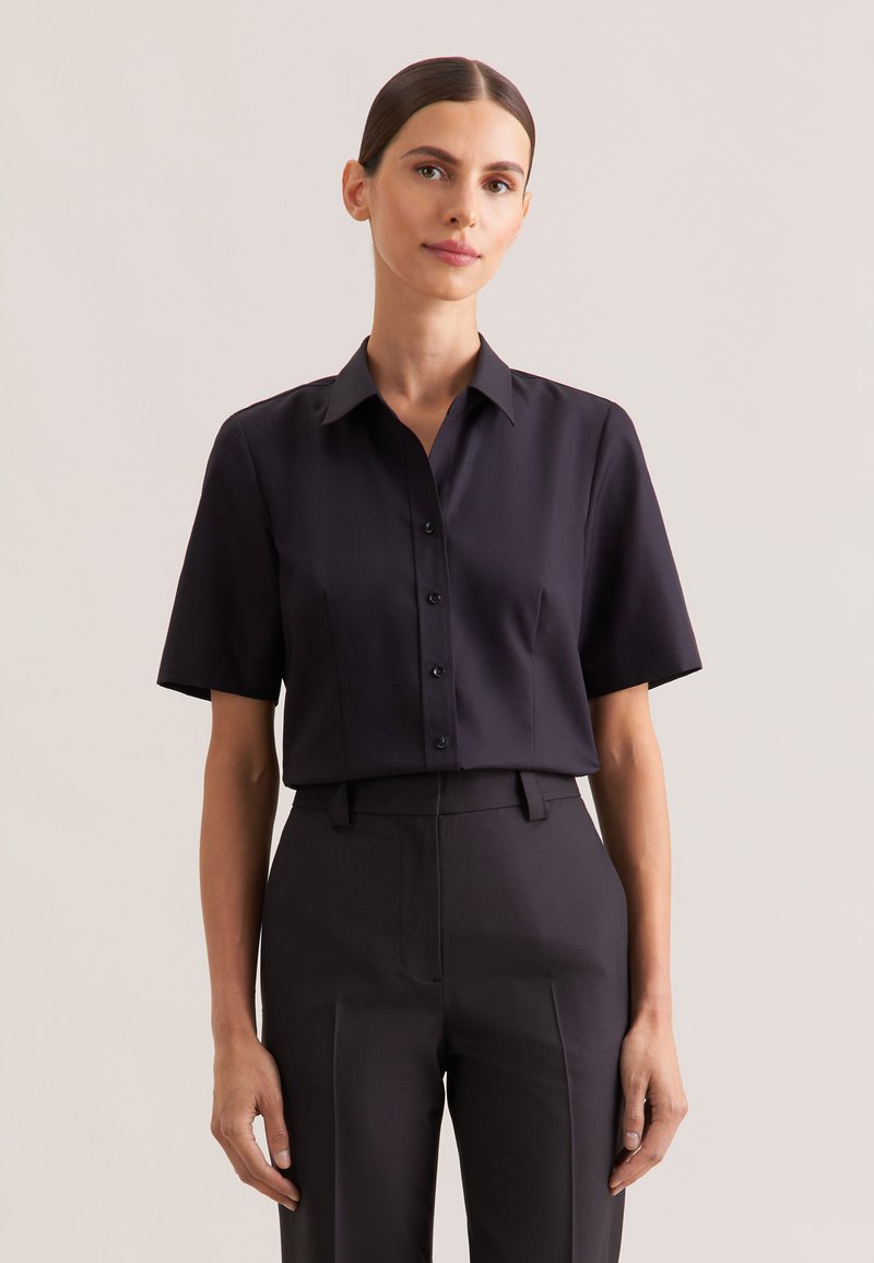 Bluzka z krótkim rękawem i kołnierzykiem w ciemnogranatowej tkaninie o gładkiej fakturze, z zapięciem na guziki z przodu i dopasowanym krojem, zestawiona z ciemnymi spodniami.
