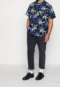 Chemise noire à manches courtes avec un motif floral coloré, associée à un jean gris retroussé et des chaussures noires à lacets. Chaussettes blanches visibles.
