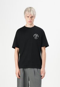 Schwarzes Baumwoll-T-Shirt mit kurzen Ärmeln, das ein weißes "Cortado Coffee"-Grafikdesign auf der linken Brust hat. Kombiniert mit grauen, gefalteten Hosen.