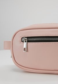 Pochette en cuir rose clair avec bords arrondis et une fermeture éclair noire horizontale équipée d'un tirette argenté sur un fond gris.