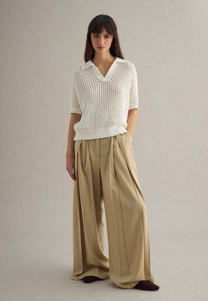 Woman standing against plain background wearing a white knit short-sleeve top and wide-leg beige trousers with hands in pockets.