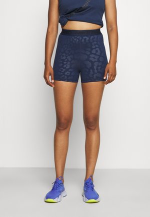 Femme portant un short de sport bleu marine à motifs, un crop top bleu marine noué et des chaussures de course bleu vif avec des semelles jaunes.