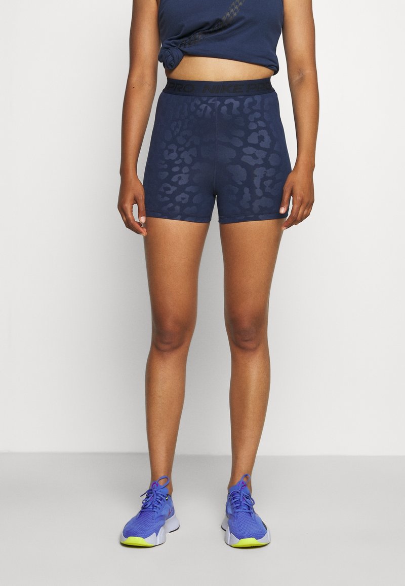Femme portant un short de sport bleu marine à motifs, un crop top bleu marine noué et des chaussures de course bleu vif avec des semelles jaunes.