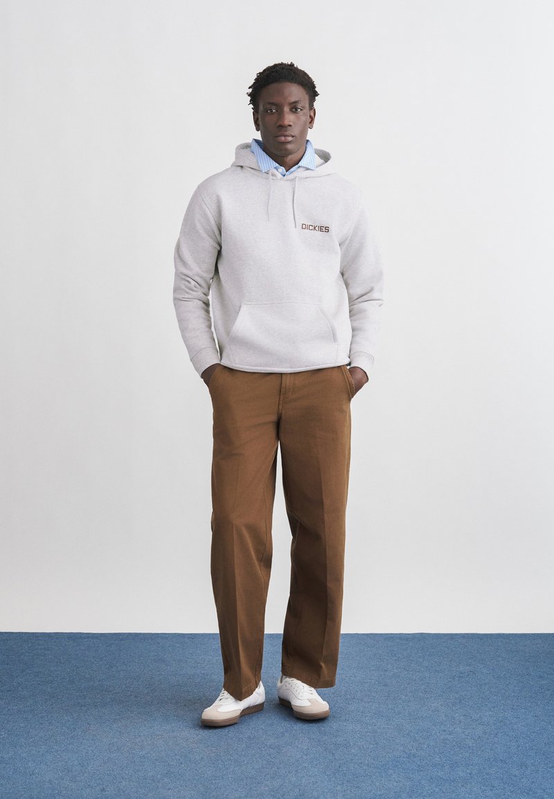 Uomo in piedi con le mani nelle tasche che indossa una felpa con cappuccio grigio chiaro con il logo "Dickies", camicia blu con colletto, pantaloni marroni e sneaker bianche su pavimento blu.