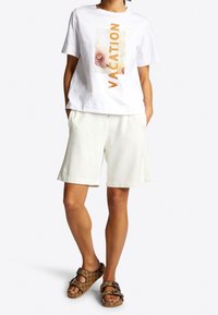 Witte katoenen T-shirt met "VACATION" print, gecombineerd met crème shorts en bruine gestreepte sandalen met gespen. Casual outfit die geschikt is voor de zomer.