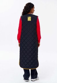 Žena s kudrnatými vlasy, oblečená v dlouhé černé prošívané vestě se žlutými detaily, pod kterou má červený svetr a černé kalhoty, stojí zády.