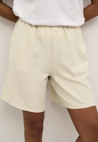 Shorts beiges texturés avec une taille élastique et des poches latérales. Le tissu semble doux, léger et conçu dans un style décontracté.