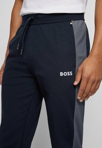 Marinblå joggingbyxor med grå sidopaneler, med dragsko i midjan och broderad "BOSS" logotyp på vänster framsida. Mjuk tygtextur.
