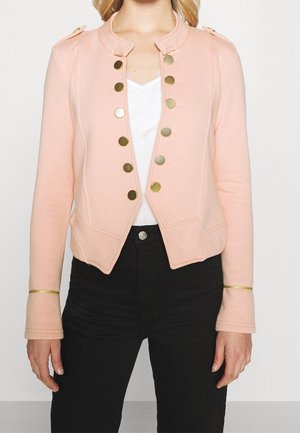 Femme portant une veste courte pêche avec des boutons dorés par-dessus un haut blanc et un pantalon noir, debout devant un fond uni.