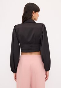 Schwarze verkürzte Jacke mit Puffärmeln, die über eine glatte Textur und Taillenbetonung verfügt. Kombiniert mit hellrosa weit geschnittenen Hosen.