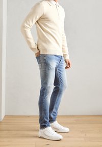 Jasno beżowy sweter z dzianiny z zamkiem przy dekolcie, zestawiony z jasnoniebieskimi dżinsami i białymi sneakersami, na neutralnym tle.