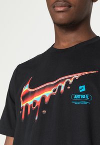 Musta puuvillapaita, jossa on värikäs, tippuva Nike-swoosh-logo ja "JUST DO IT." -teksti lihavalla sinisellä fontilla edessä.