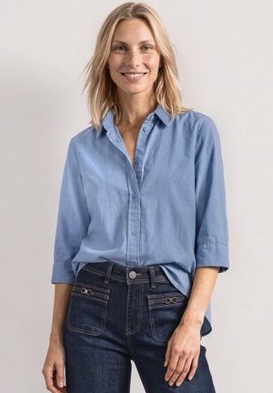 Smilende kvinde med skulderlangt blondt hår iført en lyseblå skjorte med knapper og opkørte ærmer samt mørkeblå jeans med forlommer.