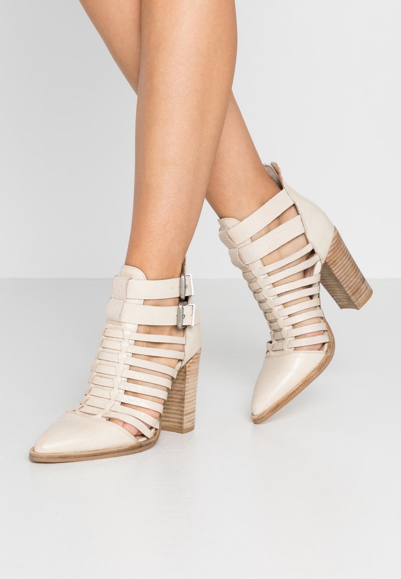 Bottes beiges à brides à la cheville avec un bout pointu, cuir texturé et talon en bois empilé. Comprend deux boucles argentées sur les côtés.