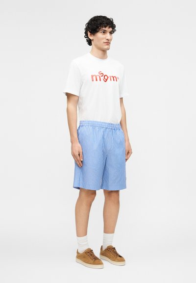 Νεαρός άνδρας όρθιος, φοράει λευκό T-shirt με σχέδιο, μπλε και λευκές ριγέ βερμούδες, λευκές κάλτσες και καφέ σουέντ αθλητικά παπούτσια μπροστά σε λευκό φόντο.