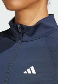 Mörkblå atletisk jacka med hög krage, dragkedja och texturerade axelpaneler. Har en vit Adidas-logotyp på nedre vänstra sidan.