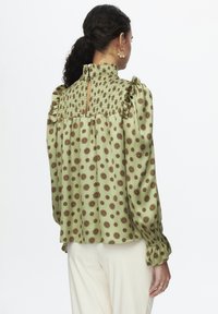 Blusa verde con design floreale a motivi, maniche arricciate, busto smockato e chiusura con bottone sul retro. Texture liscia.