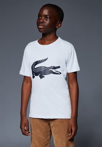 T-shirt en coton bleu clair avec un grand graphique de crocodile noir. Manches courtes et design à col ras du cou, associé à un pantalon en velours côtelé marron.